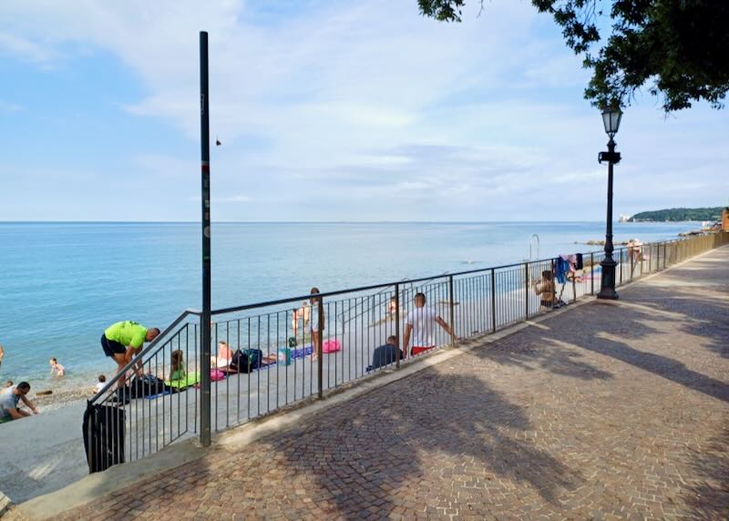 A small group of people sit on a sidewalk next to the ocean.