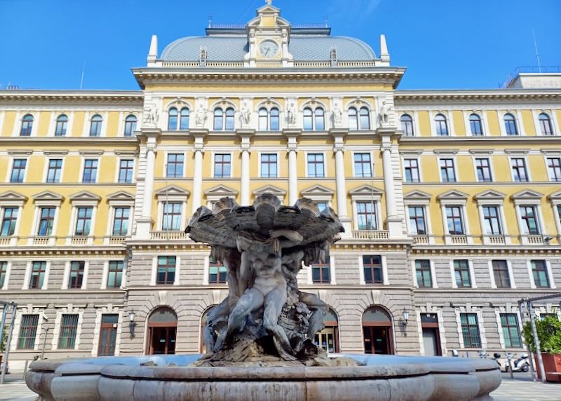 A large yellow building with a sculpture fountain in front.