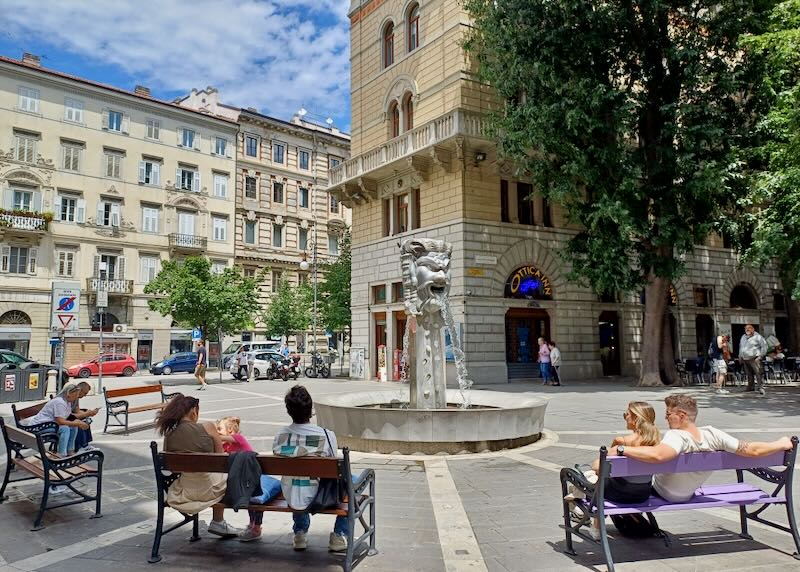 People sit on benches by a fountain on a street.